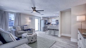 A modern living room with a grey sofa, a round ottoman, and a ceiling fan. - Recover by the Seas at Solace East - prvt pool (Holmes Beach)