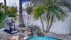 A backyard with a swimming pool, a barbecue grill, a small waterfall, and palm trees. - Recover by the Seas at Solace East - prvt pool (Holmes Beach)