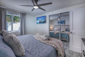 Een slaapkamer met een bed, een plafondventilator, een groot raam met gordijnen en een spiegel.
