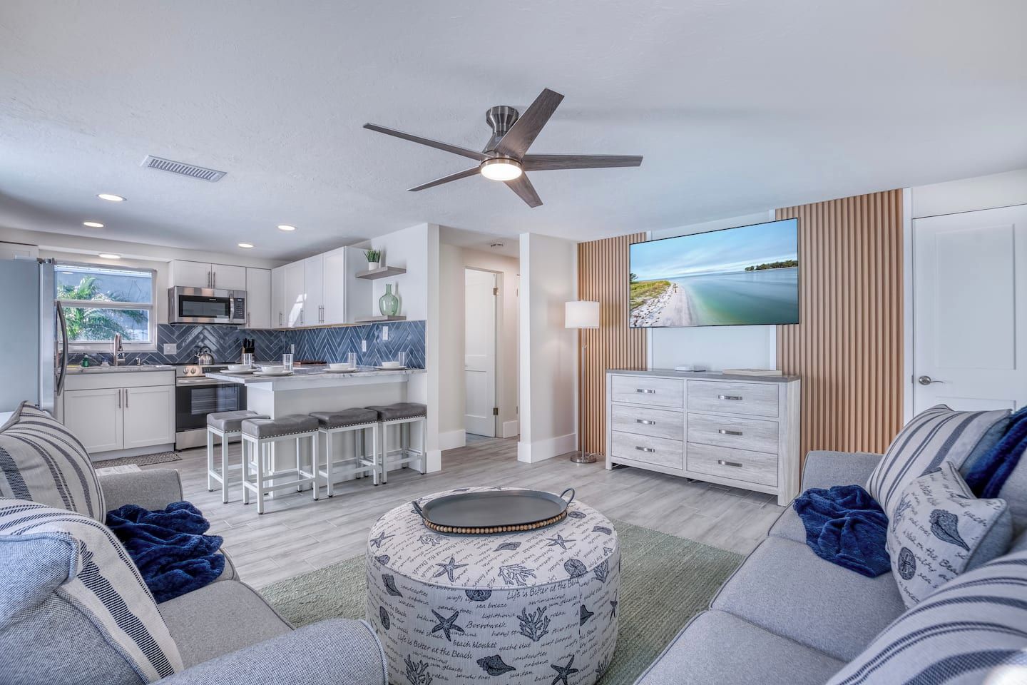 A modern kitchen with a microwave, stove, and bar stools. There is a dining area with a table and chairs, and a living room with a sofa and a television.