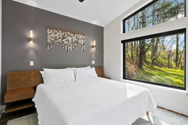 Une chambre moderne avec un grand lit, deux lampes fixées au mur, un élément décoratif mural et une grande fenêtre donnant sur une forêt.