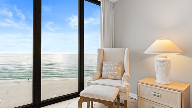 Een kamer met een groot raam met uitzicht op een strand, een fauteuil met kussen, een nachtkastje met lamp en een nachtkastje met lades.