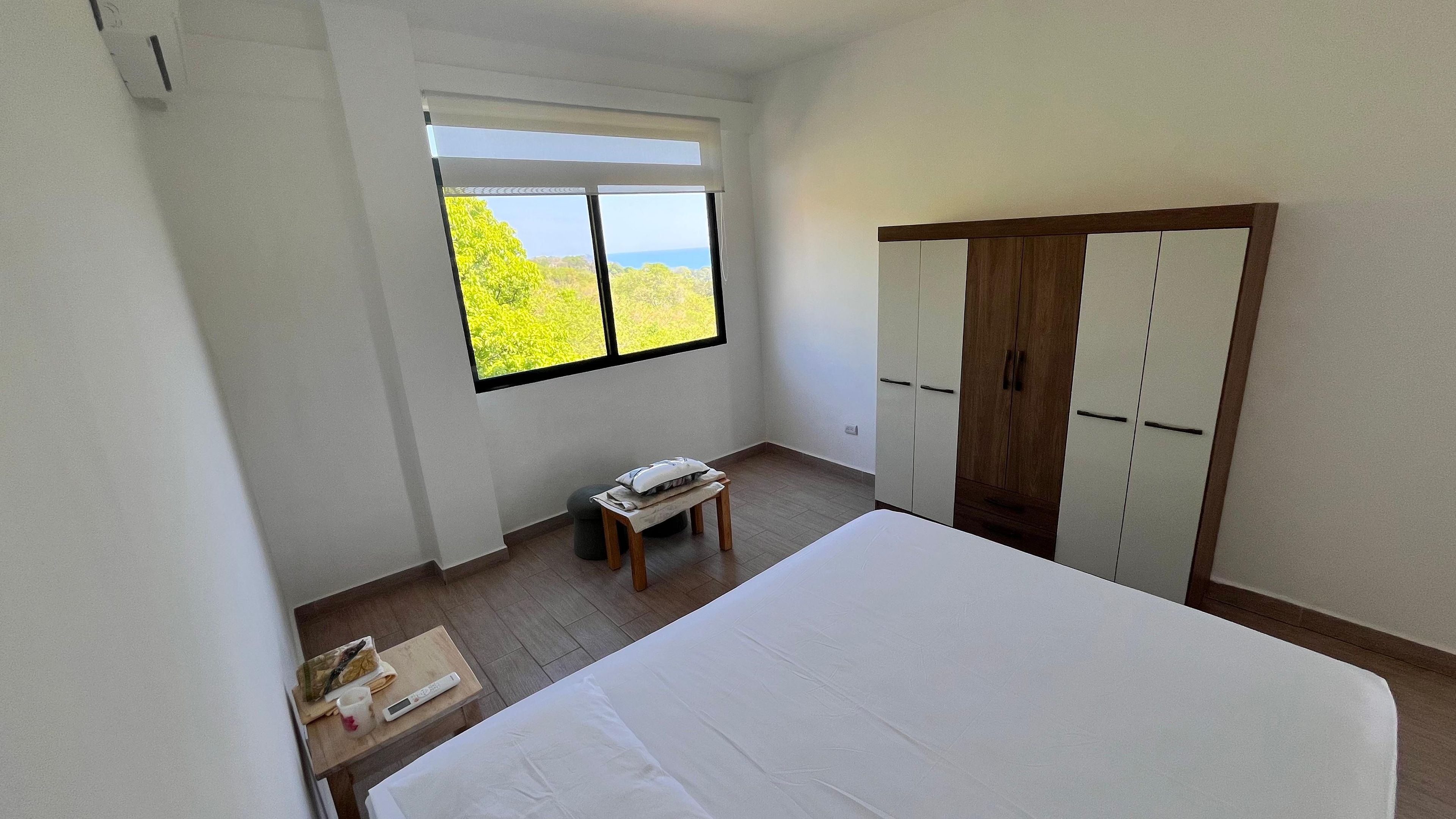 Um quarto com uma cama, janela com vista, guarda-roupa e uma pequena mesa com objetos.