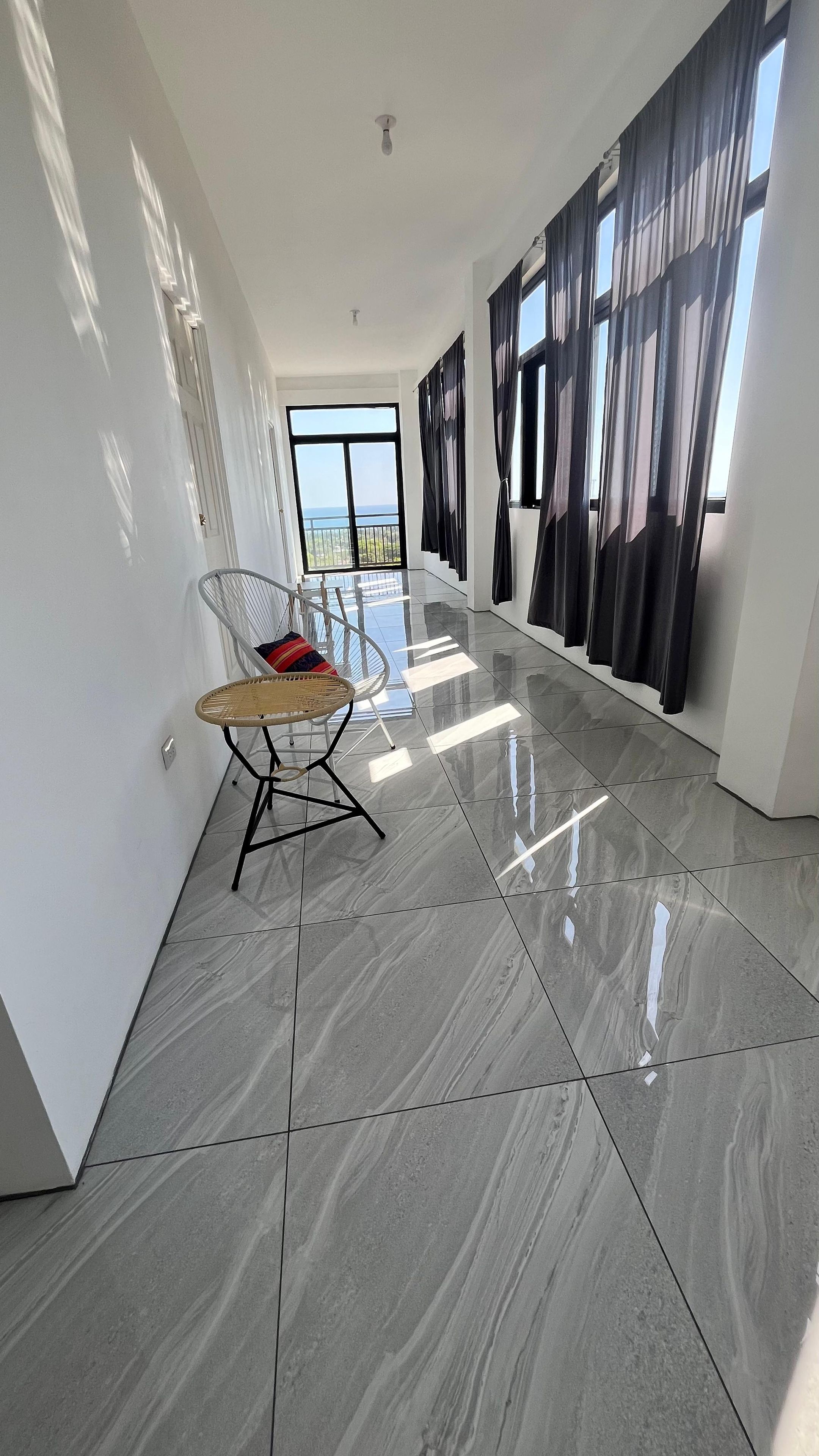 A spacious hallway with a modern design, featuring a glossy tiled floor, a wicker chair, and a large window with curtains.