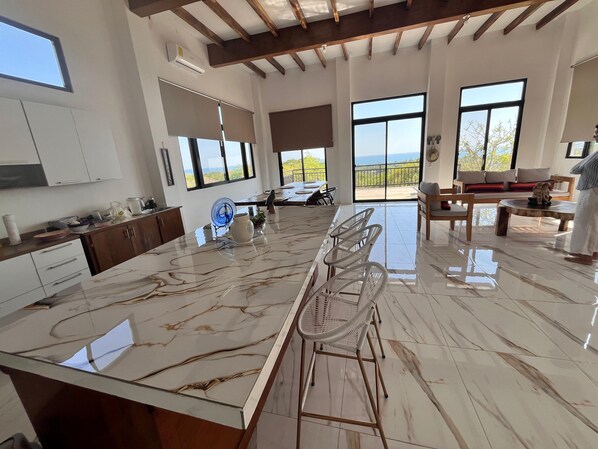 A modern kitchen with a marble countertop, a dining area with chairs, and large windows offering a view.