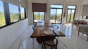 Une pièce spacieuse avec une grande table à manger en bois, des chaises noires et une vue sur l’extérieur grâce à de grandes fenêtres.