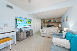 Een woonkamer met een piano, een tv waarop een strandscène te zien is, een bank en een stenen muur met decoratieve objecten.