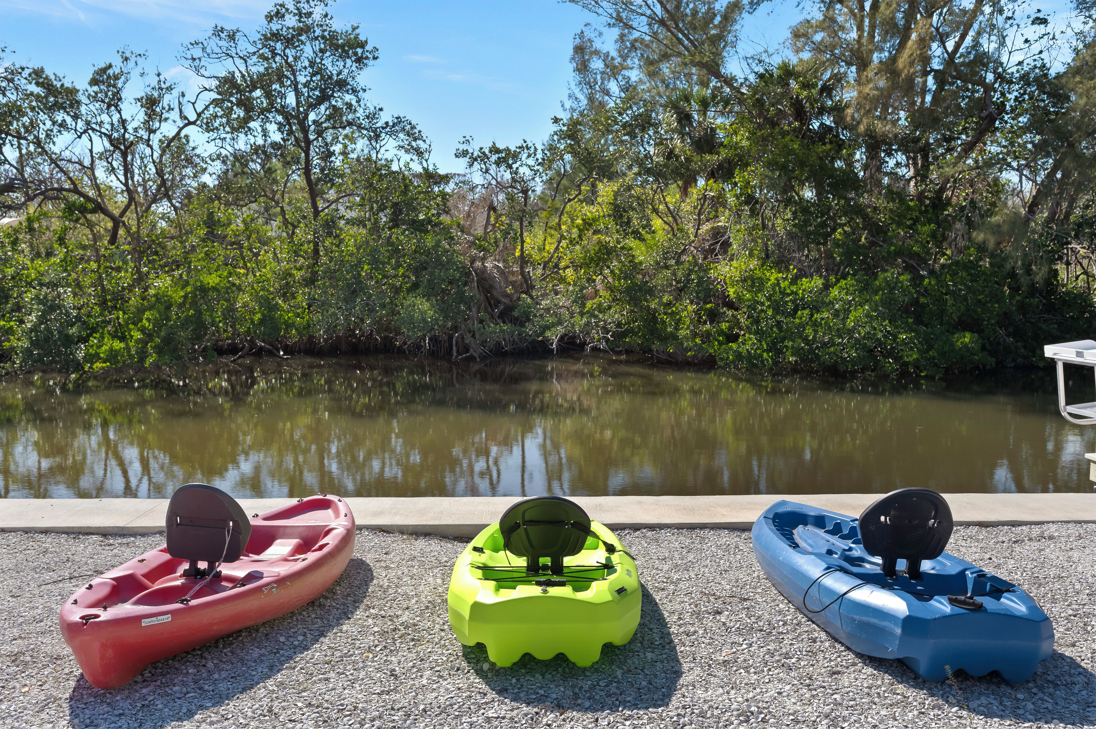 Tres kayaks alineados en un camino de grava junto a una masa de agua tranquila, rodeados de abundante vegetación.