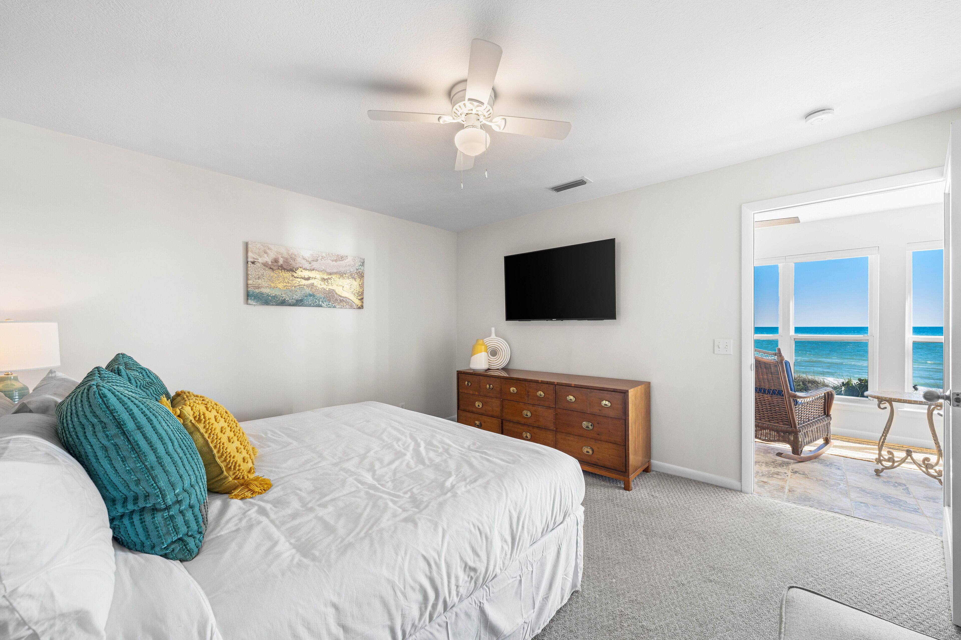 A bedroom with a bed, a dresser, a ceiling fan, a TV, and a view of the ocean through a door.