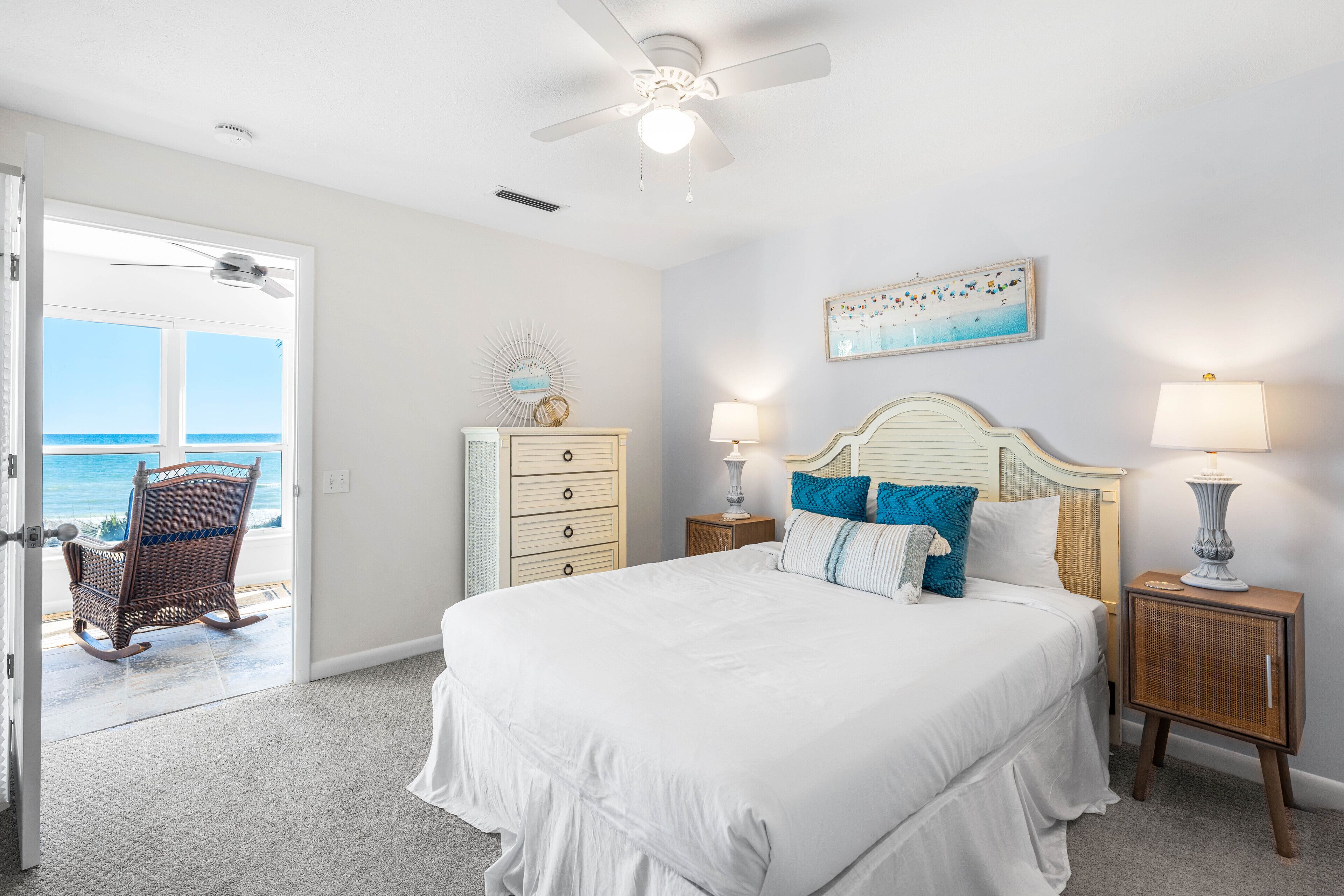 A bedroom with a large bed, two bedside lamps, a ceiling fan, a dresser, and a rocking chair near a door leading to a balcony with a view of the ocean.