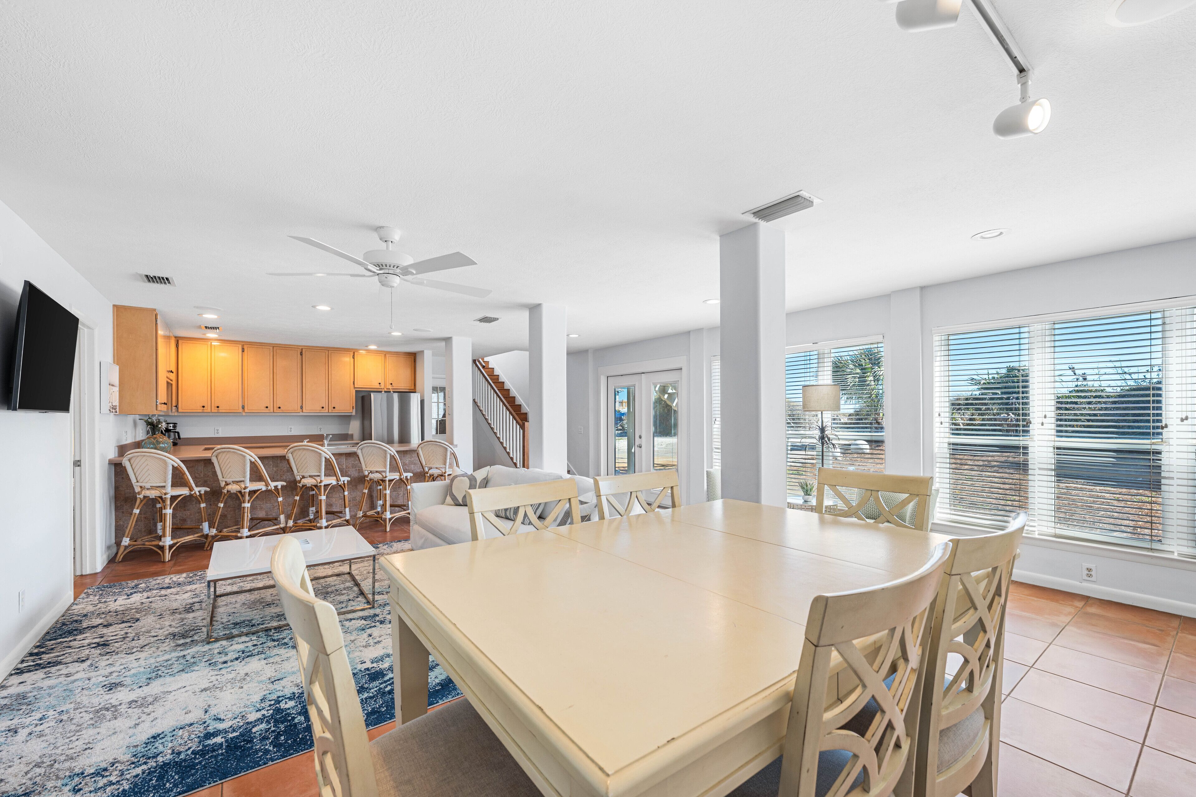 A spacious dining area with a large table and chairs, a kitchen in the background, and large windows with blinds.