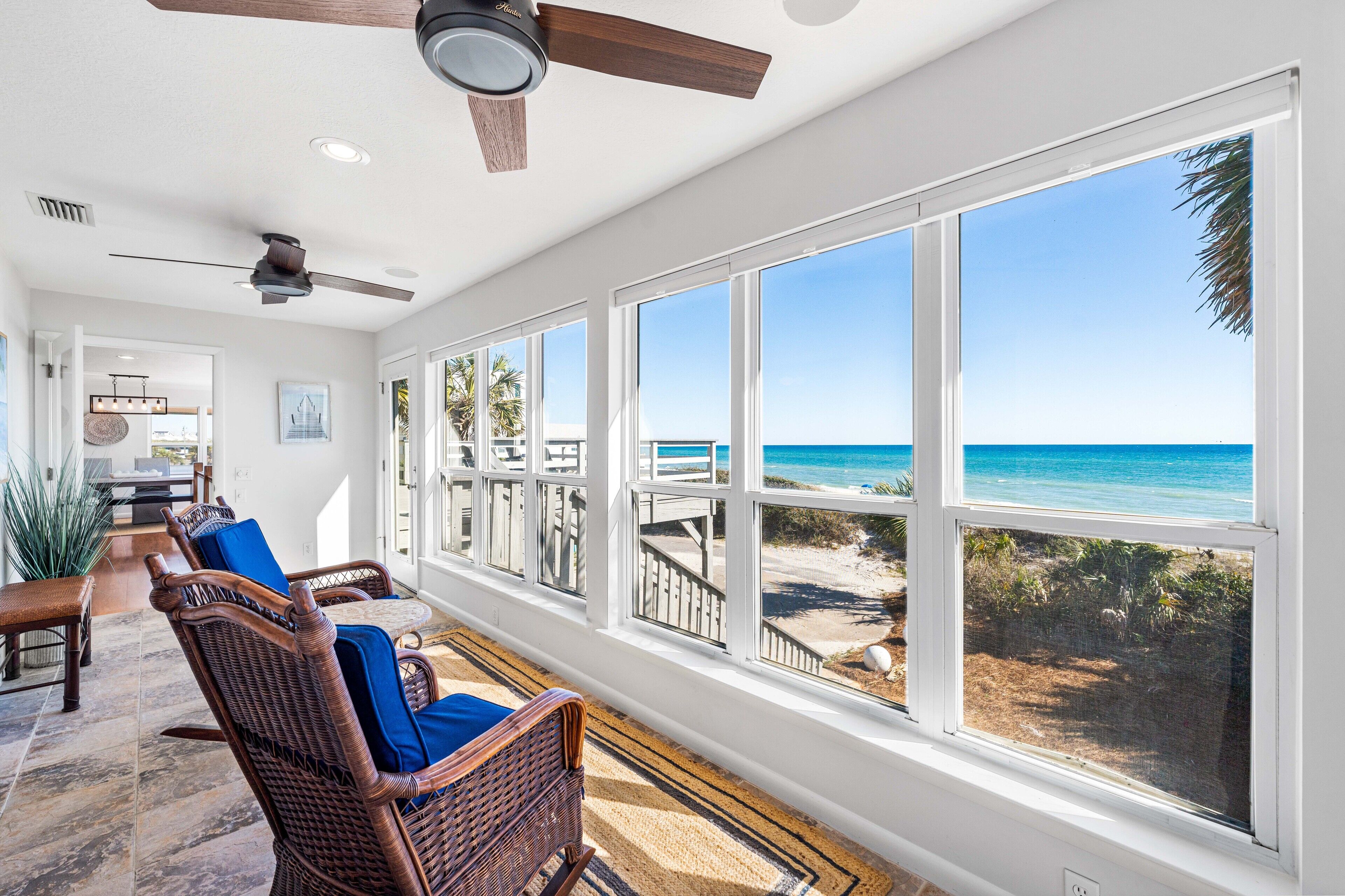 A room with large windows overlooking a beach, wicker furniture, and ceiling fans.