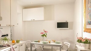 A modern dining area with a white table set for four, a flat-screen TV mounted on the wall, and a potted plant with pink flowers on the right. - Riccardo Condotti Suite (Rome)