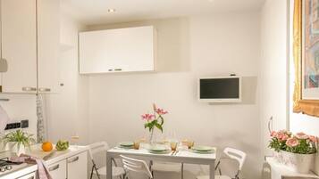 A modern dining area with a white table set for four, a flat-screen TV mounted on the wall, and a potted plant with pink flowers on the right.