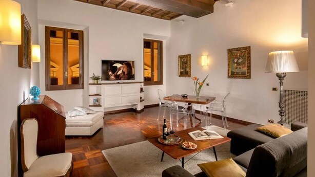 A modern living room with a wooden ceiling, a white sofa, a dining table with chairs, and a television. - Riccardo Condotti Suite (Rome)
