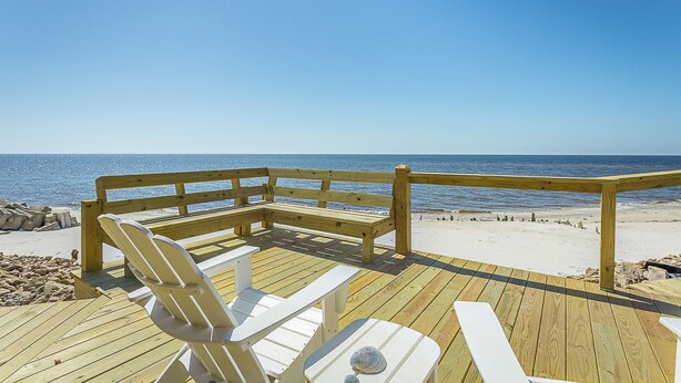 Una terraza de madera con banco y sillas, con vistas a una playa bajo un cielo azul y despejado.