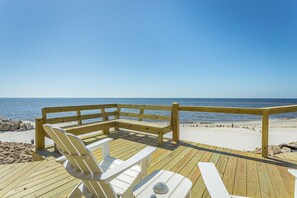 A wooden deck with a bench and chairs overlooking a beach with a clear blue sky.