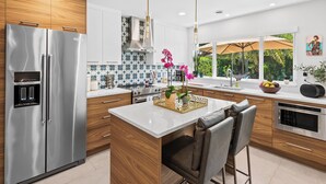 Een moderne keuken met een roestvrijstalen koelkast, houten kastjes, een wit aanrecht en een groot raam met uitzicht op groen.