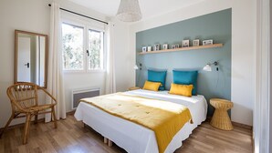 A bedroom with a bed, a chair, a bedside table, a shelf with pictures, and a window. - Villas Indigo, Villa(s) Indigo proches des plages (La Teste-de-Buch)
