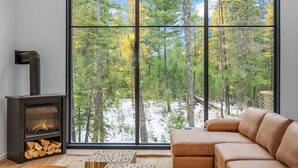 A cozy living room with a large window, a fireplace, and a brown leather sofa. - Off Grid Stays - Treehouse #1 (Columbia Falls)