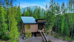 A cabin with a metal roof, a wooden deck, and a staircase leading to a balcony. - Off Grid Stays - Treehouse #1 (Columbia Falls)