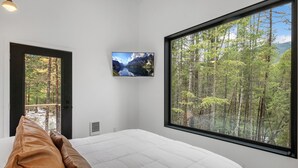 A bedroom with a large window offering a forest view, a bed with white bedding, and a mounted picture of a mountain lake. - Off Grid Stays - Treehouse #1 (Columbia Falls)