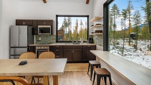 A modern kitchen with dark wood cabinets, a stainless steel refrigerator, and a large window offering a view of a snowy forest. - Off Grid Stays - Treehouse #1 (Columbia Falls)