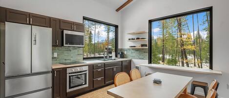 Una cocina moderna con armarios oscuros, electrodomésticos de acero inoxidable y un amplio ventanal con vistas a un bosque.