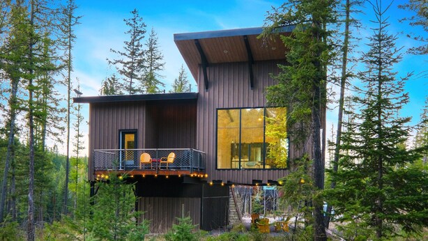 A modern cabin with a balcony, large windows, and a forest backdrop. - Off Grid Stays - Treehouse #1 (Columbia Falls)
