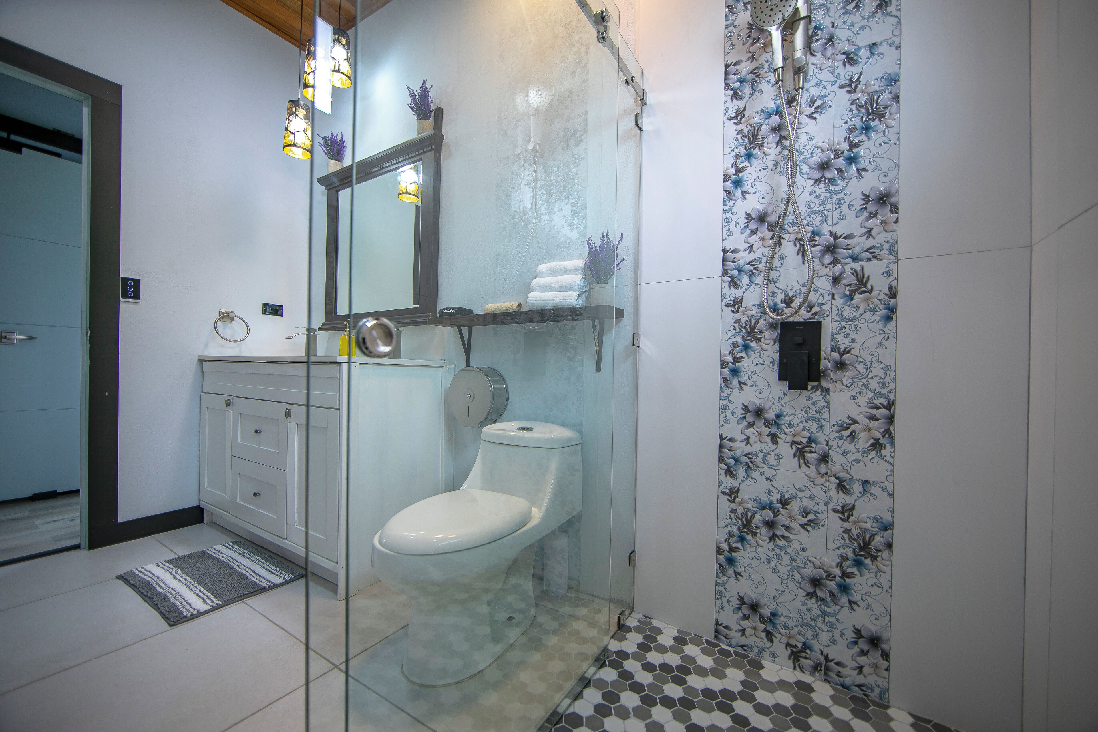 Een badkamer met een glazen douchecabine, een toilet, een wastafel met spiegel en een betegelde spatwand met een patroon.