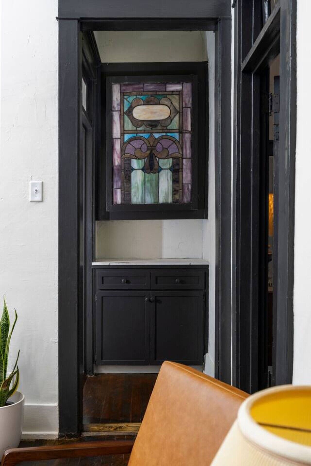 A narrow hallway with a stained glass window, a dark wooden door, and a white wall.