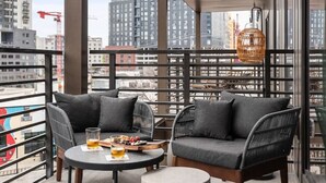 A balcony with two sofas, a round table, and a small stool. The balcony has a metal railing and large windows offering a city view.