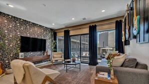 Una sala de estar moderna con un televisor grande, asientos cómodos, una mesa de centro y vistas a la ciudad a través de amplias ventanas.
