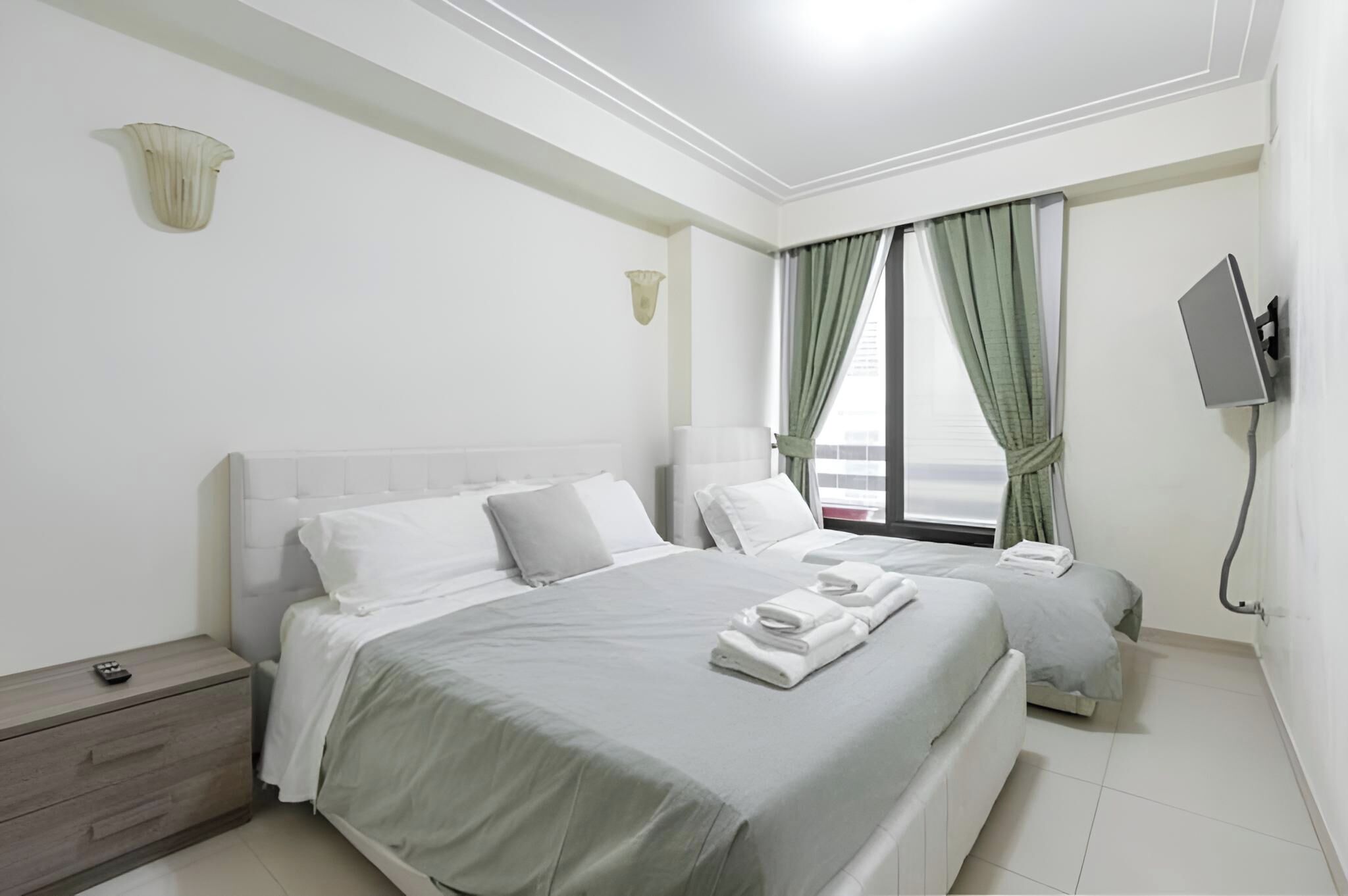 Une chambre d’hôtel avec un grand lit, un lit plus petit, un téléviseur fixé au mur et une fenêtre avec des rideaux.