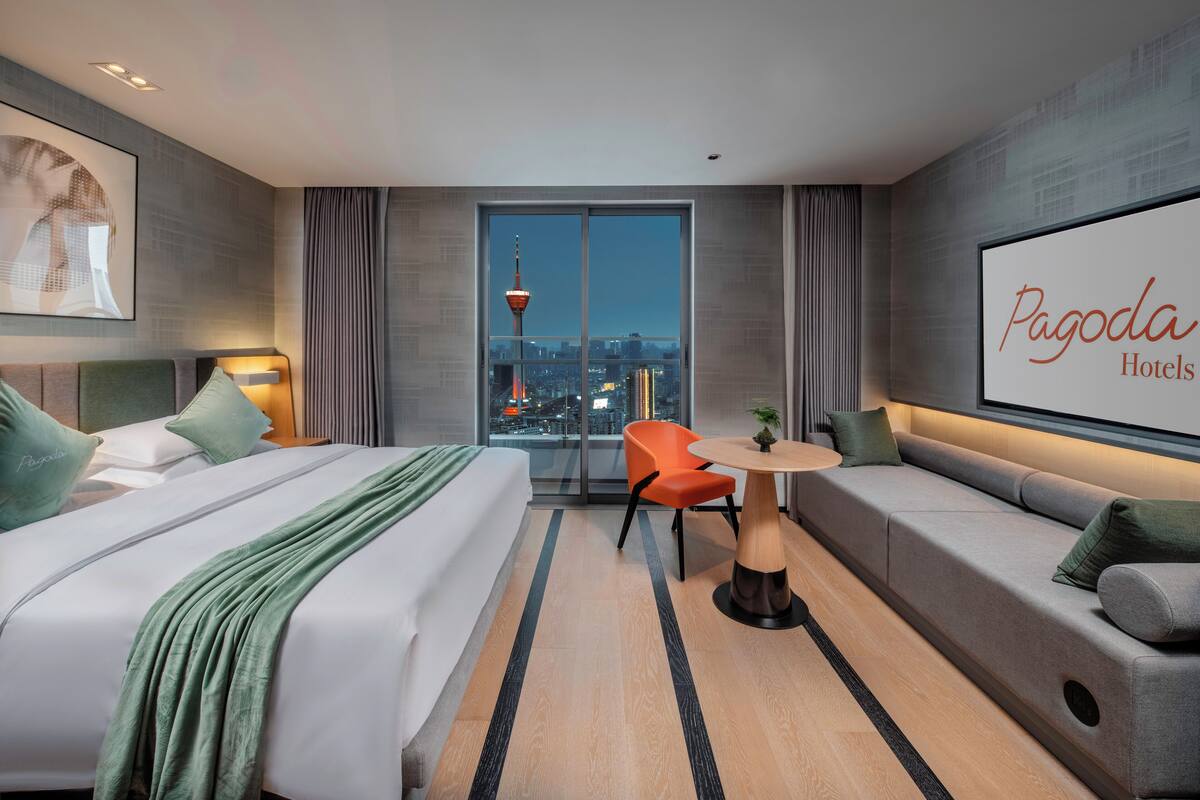 A modern hotel room with a large bed, a sofa, a small table, and a city view.