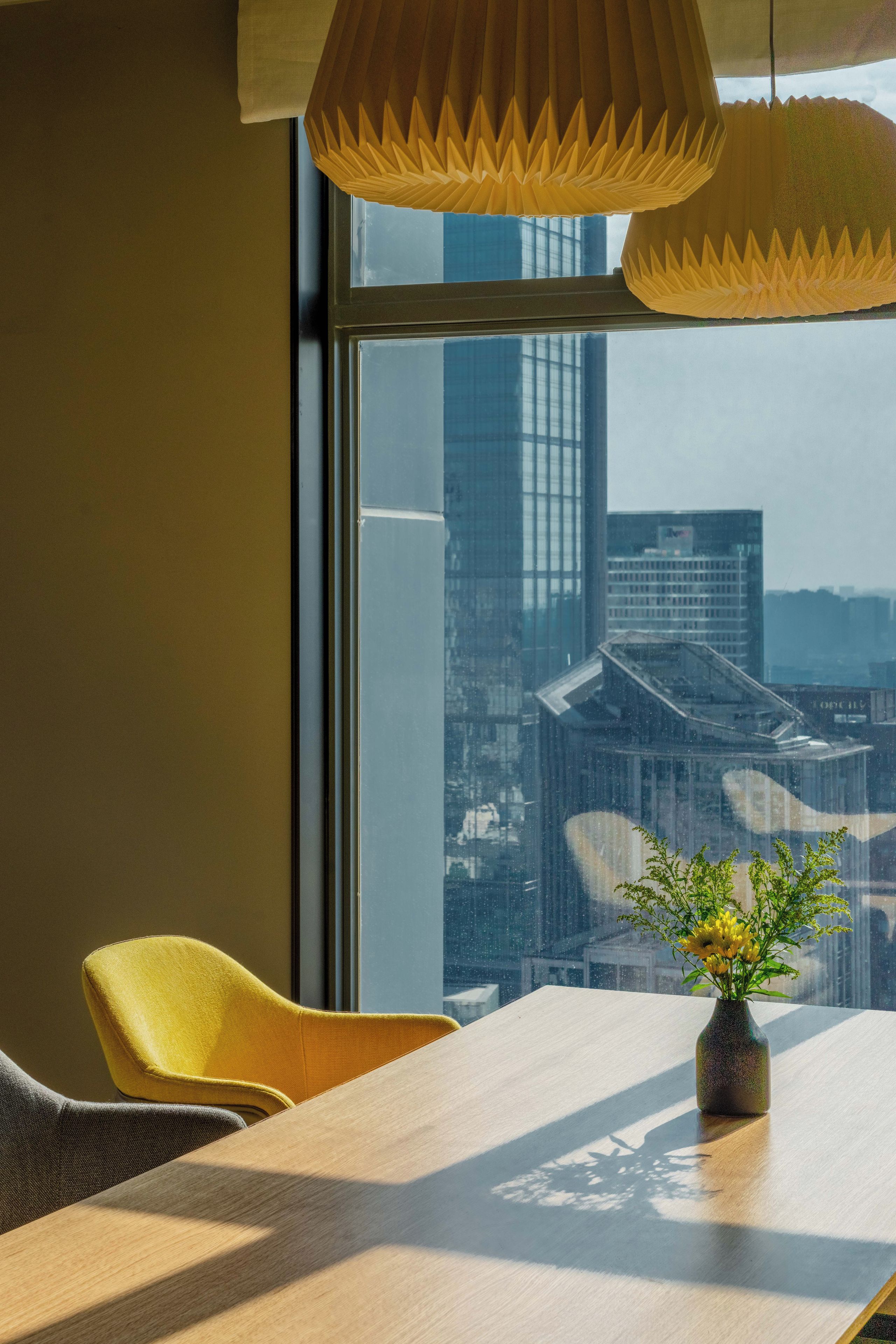 A modern interior with a dining table, chairs, a vase with flowers, and a view of a cityscape through a large window.