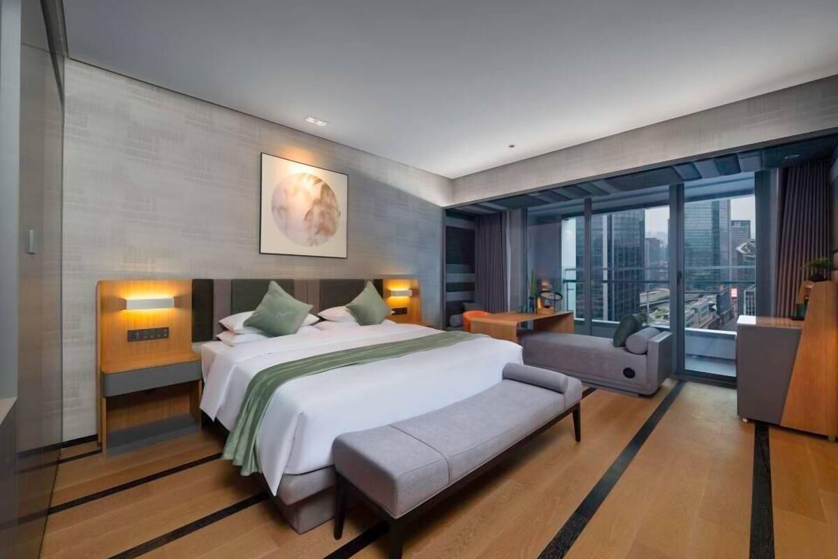 A modern hotel room with a large bed, a bench, a desk, and a view of the city through large windows.