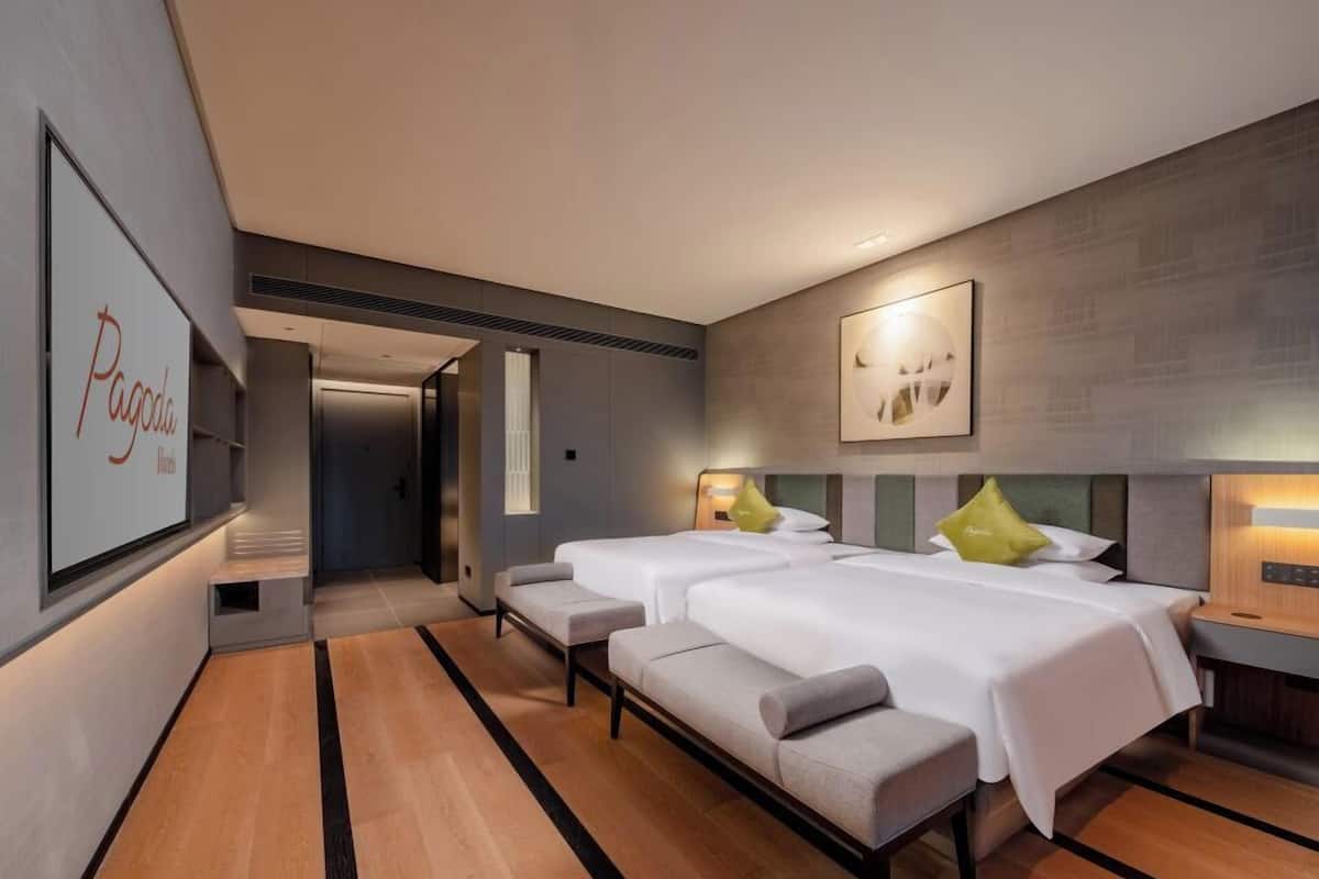 A modern hotel room with two beds, a bench, a wall-mounted TV, and a door leading to another room.