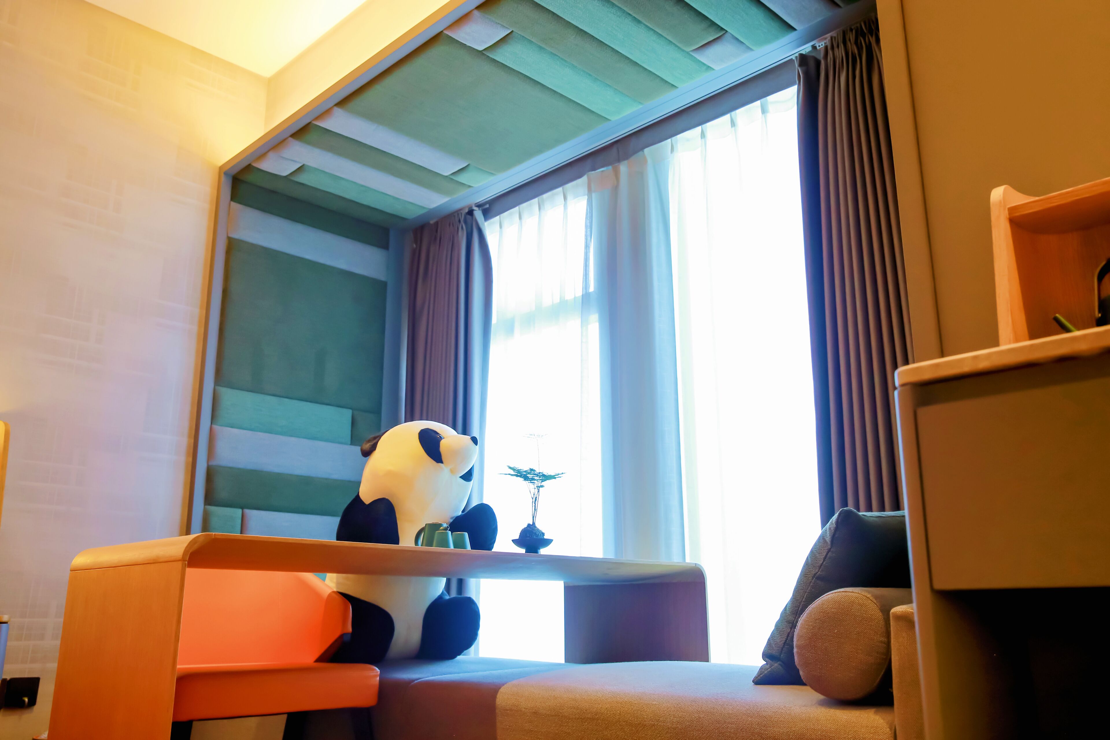 Une chambre avec un lit, une fenêtre avec des rideaux, une petite table avec une plante et un grand ours en peluche à l’effigie d’un panda.