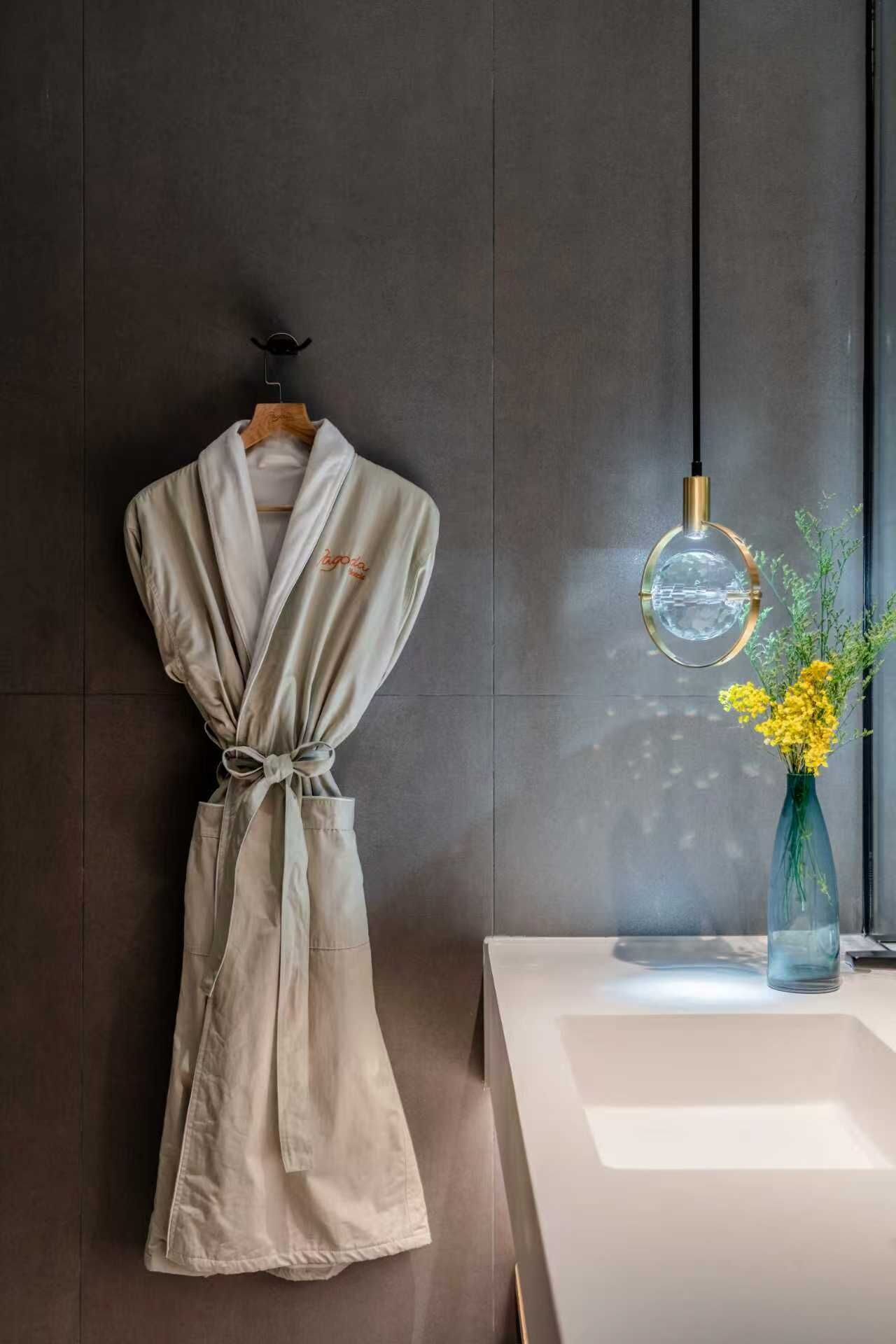 A modern bathroom with a robe hanging on a wooden hanger, a round mirror, and a vase with yellow flowers on a white sink.