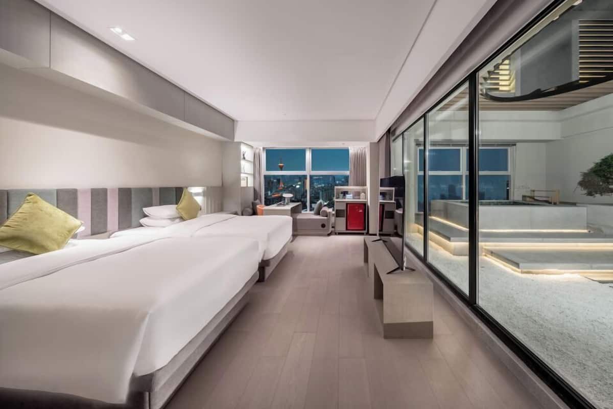 A modern hotel room with a large bed, a desk, and a view of the cityscape through floor-to-ceiling windows.