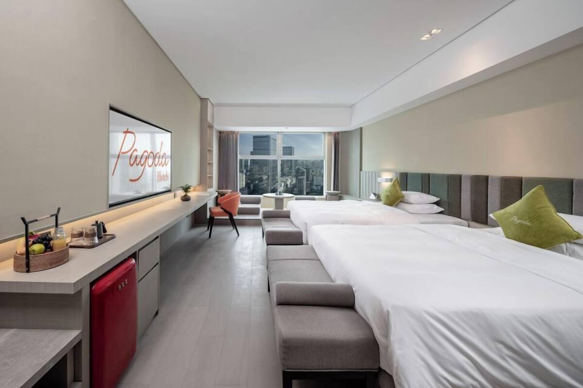 A modern hotel room with a large bed, a desk with a TV, a red fridge, and a city view.