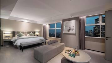 Quarto de hotel moderno com uma cama grande, um sofá, uma mesa de jantar e vista para a cidade.
