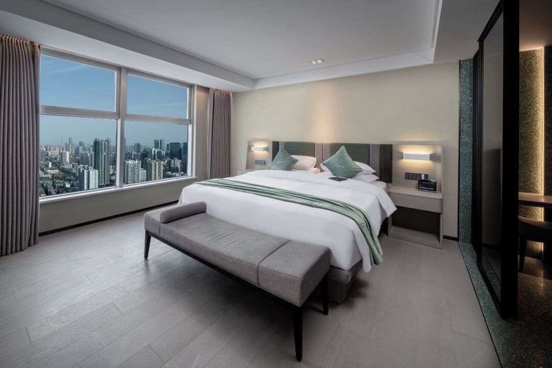 A modern hotel room with a large bed, a bench, a nightstand, and a view of the city.