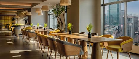 A modern dining area with wooden tables and chairs, large windows with a city view, and hanging pendant lights.