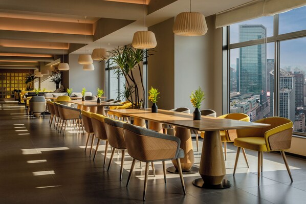 Un comedor moderno con mesas y sillas de madera, ventanas amplias con vista a la ciudad y lámparas colgantes.