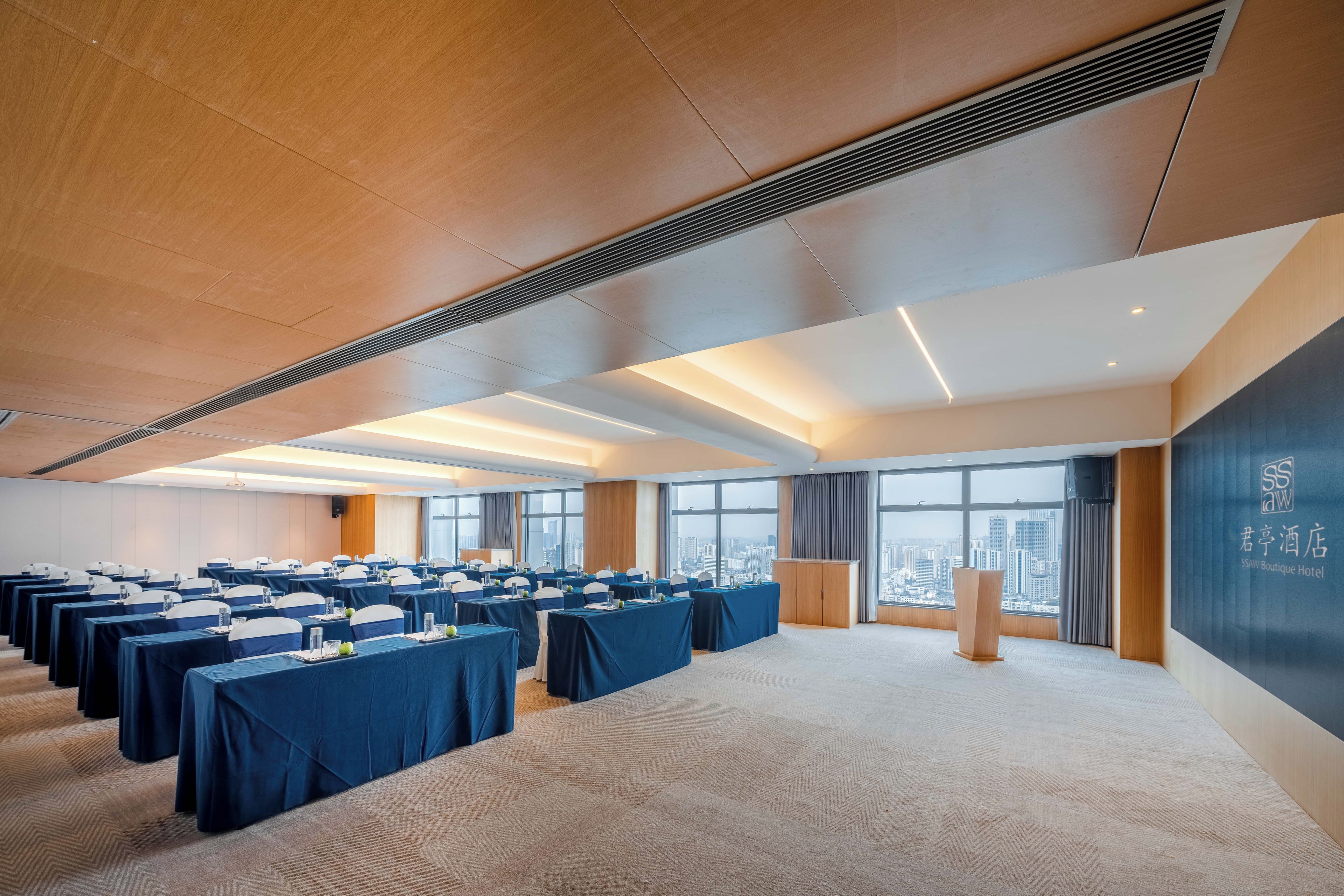 Un salón de conferencias con varias mesas, un atril y vistas a la ciudad a través de amplias ventanas.