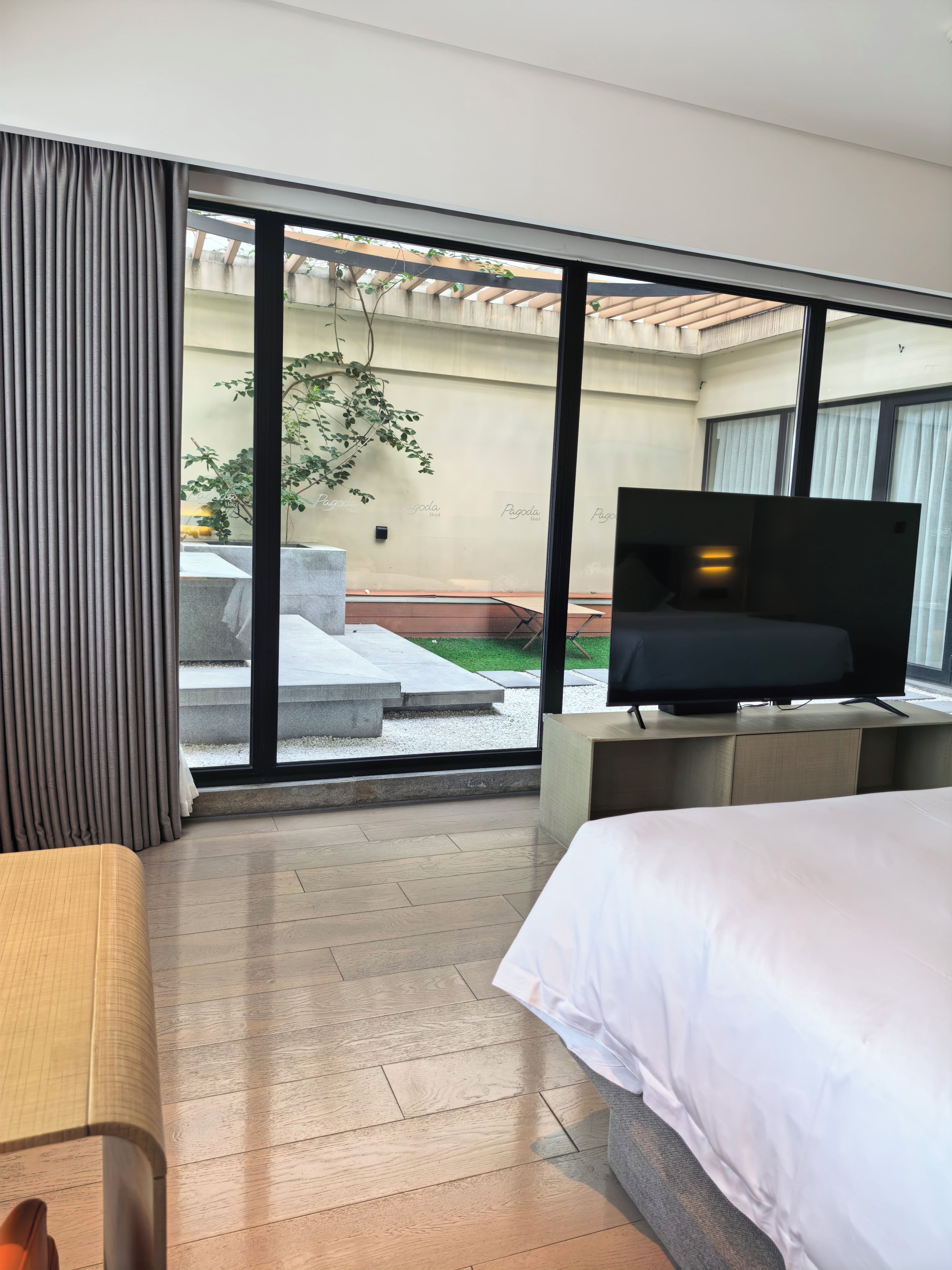Habitación de hotel con cama, televisor sobre un mueble y vistas a un espacio exterior con escaleras y vegetación.