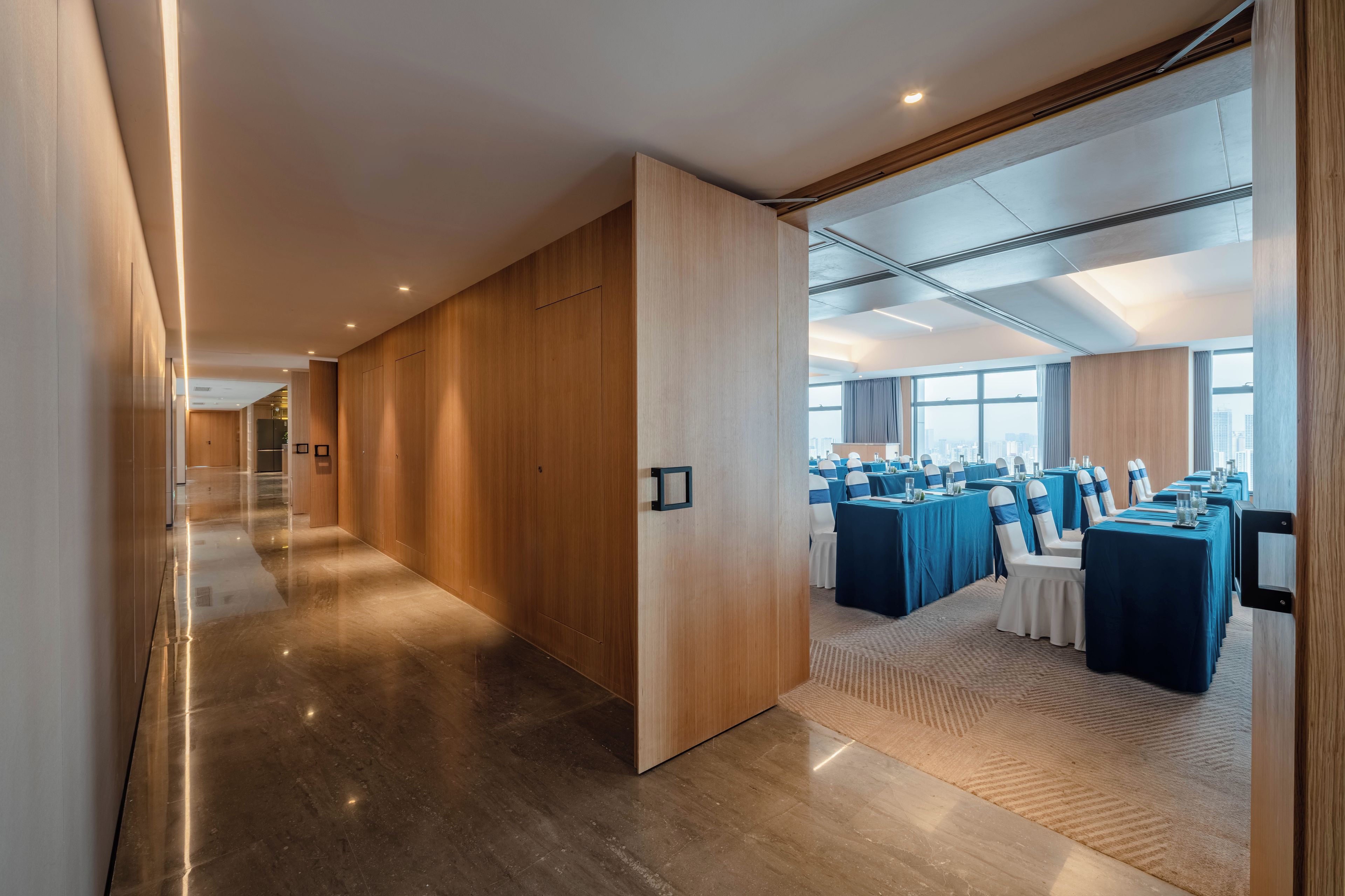 A modern hotel corridor with wooden paneling, a conference room with tables and chairs, and a view of the city through large windows.