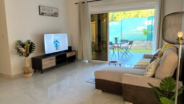 Ein modernes Wohnzimmer mit Flachbildfernseher, einer Couch, einem Couchtisch und einem Essbereich, der durch Glastüren sichtbar ist.